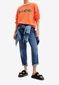 Oranje sweatshirt met de tekst "POWER", spijkerbroek geknoopt om de taille en zwarte sandalen. Casual, relaxte pasvorm met opgerolde mouwen van de broek.
