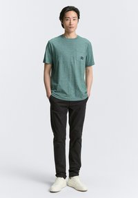 Groene gestreepte t-shirt met korte mouwen, ronde halslijn en zakje op de linker borst, gecombineerd met zwarte broek en witte schoenen.