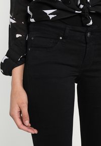 Jeans de mezclilla negra con un corte ajustado, con bolsillos frontales, cierre de botón y un detalle de costura minimalista, combinados con una blusa estampada.