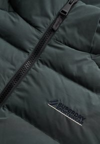 Ciemnozielona kurtka pikowana z czarnym zamkiem błyskawicznym oraz wyszywanym logo "Superdry Sportswear" w granatowym i białym kolorze na materiale.