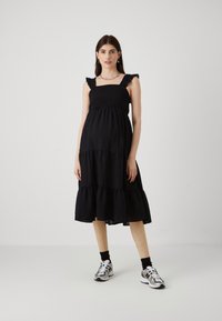 Rochie neagră cu straturi, cu bretele volanase, bust texturat și țesătură fluidă. Asortată cu adidași sport și șosete până la gleznă pentru contrast.