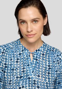 Blaue Bluse mit weißen Punkten, gemustertem Stoff, Knoten am Hals und weicher Textur. Die Bluse hat geraffte Schultern für mehr Volumen.