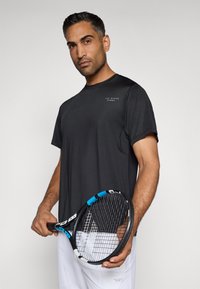 Čierne športové tričko s krátkymi rukávmi a okrúhlym výstrihom, s bielym logom. Model drží tenisovú raketu s modrým akcentom.