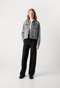 Grijze denimjack met knoopsluiting, twee borstzakken en een relaxte pasvorm; gedragen over een witte shirt en zwarte wijde broek.
