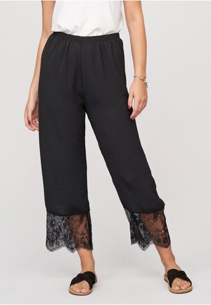 Mujer con pantalones negros cortos con bordes de encaje en los puños, una camiseta blanca, sandalias negras y una pulsera plateada en la muñeca derecha.