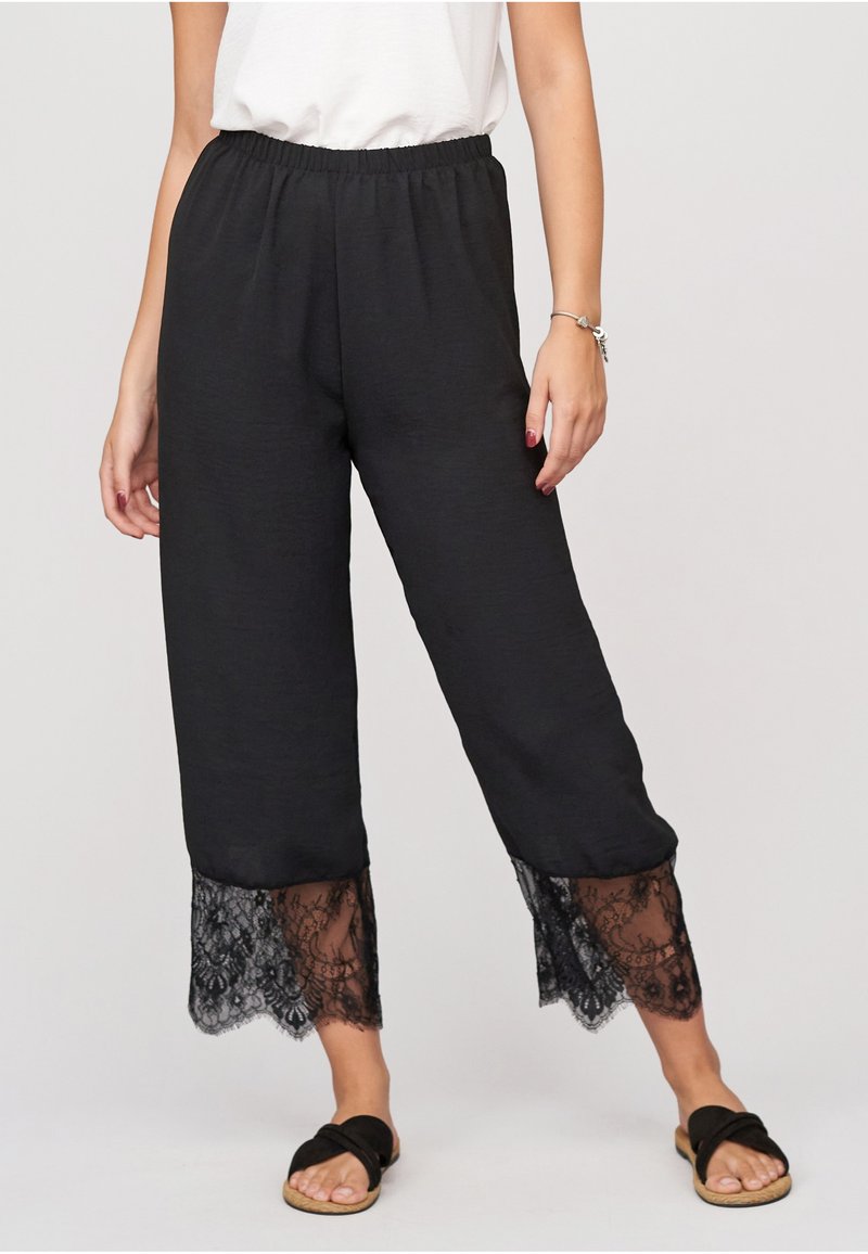 Femme portant un pantalon noir court avec bordure en dentelle aux poignets, un haut blanc, des sandales noires et un bracelet argenté au poignet droit.