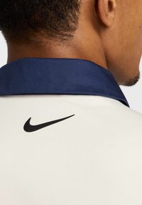 Biała koszulka polo z ciemnoniebieskim kołnierzykiem, z czarnym logo Nike na plecach. Tkanina wydaje się gładka o jednolitej fakturze.