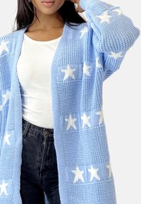 Cardigan lavorato a maglia azzurro chiaro con motivi a stelle bianche, design aperto e tessuto testurizzato. Indossato sopra una maglietta bianca a coste e pantaloni neri.