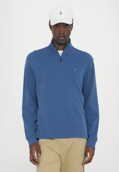 Blauwe quarter-zip pullover van zachte stof met lange mouwen en geborduurd logo, gecombineerd met een witte pet met een vergelijkbaar logo en beige broek.