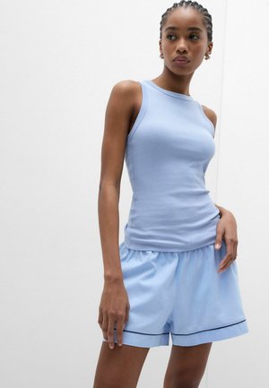 Kvinde med flettet hår iført en ærmeløs lyseblå ribbet top og løstsiddende lyseblå shorts med sort kant, stående mod en hvid baggrund.