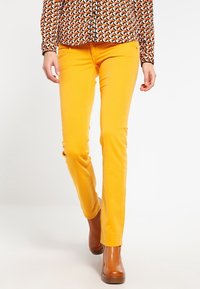 Pantalones de algodón amarillos con un corte ajustado, que presentan un estilo de cinco bolsillos, combinados con botas de tobillo de tacón marrón y una blusa de manga larga con estampado.