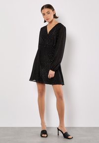 Młoda kobieta stoi w czarnej, długorawej sukience mini w wzór zygzak i czarnych, wysokich, odkrytych sandałach na obcasie na tle białym.