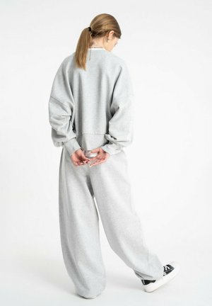 Młoda kobieta z kucykiem, ubrana w luźną szarą bluzę i szerokie szare spodnie, stoi z złączonymi za plecami rękami, jedna stopa lekko uniesiona.