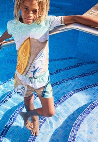 Grafisk T-shirt med tryck av turkost vatten och surfbräda; matchande badshorts med surfbrädesdesign i livliga färger. Står i en pool.