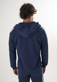 Sudadera azul marino hecha de tela suave, con capucha ajustable y rayas blancas en las mangas, combinada con pantalones a juego.