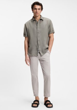 Erwachsener Mann, der ein hellolivgrünes kurzärmeliges Hemd mit Knöpfen trägt, beigefarbene Hose und schwarze Sandalen mit Schnalle, steht mit einer Hand in der Tasche.