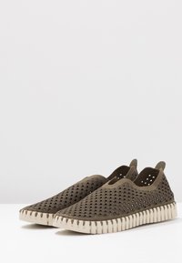 Scarpe slip-on verde oliva con una tomaia ventilata e perforata e una suola spessa, bianca e texturizzata caratterizzata da dettagli di cuciture a zigzag.