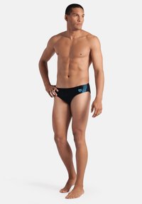 Bañador de hombre negro con detalles geométricos azules en el lado izquierdo. Material liso con un diseño ajustado. Modelo de pie descalzo con las manos en las caderas.