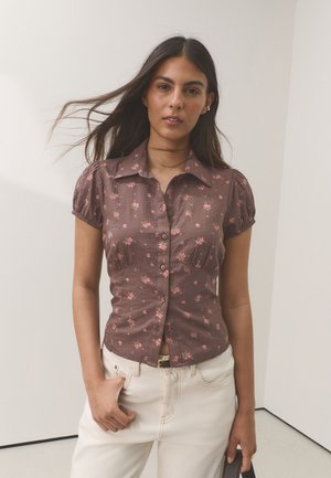 Vrouw met lang donker haar draagt een bruin, kort mouwtjes, knoophemd met roze bloemenprint en witte broek, staand in een neutrale kamer.