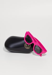 Roze sunčane naočale s crnim staklima odložene na crnoj, sjajnoj futroli koja ima okrugli zlatni logo. Oblik je kvadratičan s lagano zaobljenim rubovima.