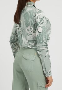Grün bedruckte Bluse mit floralem Muster, langen Ärmeln, Kragen und glatter Satin-Textur; kombiniert mit hochgeschnittenen olivgrünen Hosen mit aufgesetzten Taschen.