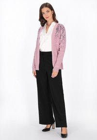 Světle růžový cardigan se zdobením z flitrů na ramenou, kombinovaný s černými širokými nohavicemi a černými podpatky. Pod ním bílá košile s knoflíky.