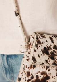 Cowhide crossbody bag featuring a spotted pattern in cream and brown, with a flat flap and knotted strap. Textured surface and soft finish.
