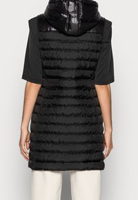 Czarna pikowana kamizelka z błyszczącym kapturem, z poziomymi przeszyciami i bez rękawów. Materiał wydaje się gładki i lekko błyszczący.