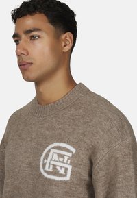 Młody mężczyzna z krótkimi kręconymi włosami, ubrany w jasnobrązowy sweter z dzianiny, ozdobiony białym geometrycznym logo na piersi, zwrócony w lewo.