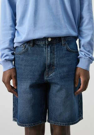Personne portant une chemise bleue à manches longues et un short en jean bleu foncé ample, debout avec les mains détendues le long du corps.