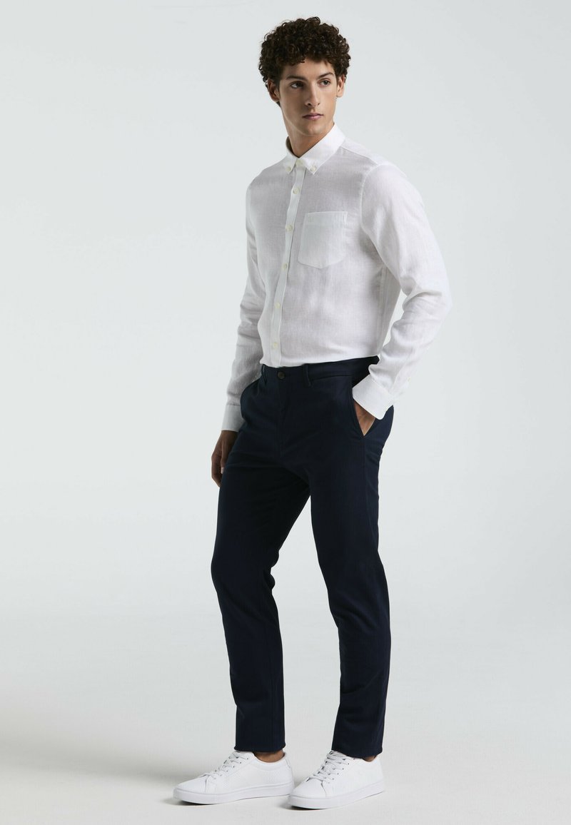 Witte linnen overhemd met knoopsluiting en een borstzak, gecombineerd met donkerblauwe op maat gemaakte broek en witte sneakers. Zachte textuur en elegante uitstraling.