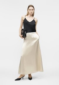 Γυναίκα που φοράει μαύρο τοπ με λεπτές τιράντες, κρεμ σατέν μακριά φούστα, μαύρα μυτερά τακούνια, κρατώντας μαύρη τσάντα, στέκεται μπροστά σε λευκό φόντο.