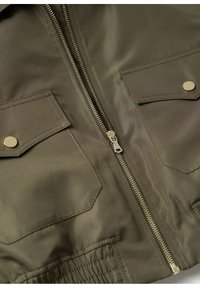 Veste vert olive avec fermeture éclair métallique centrale, deux poches à rabat avec boutons-pression dorés et ourlet élastiqué en bas.