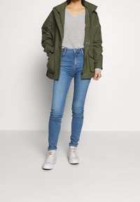 Persona che indossa una giacca verde oliva aperta su un maglione grigio a coste, jeans skinny blu e sneakers alte bianche, in piedi su uno sfondo neutro.