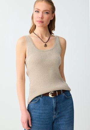 Donna che indossa un top beige senza maniche a coste, jeans blu con cintura marrone e una collana con ciondolo rotondo su cordoncino scuro.