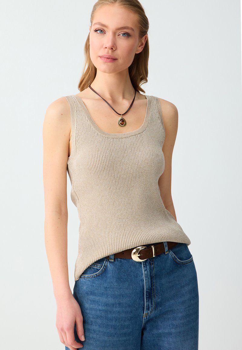 Femme portant un haut beige sans manches côtelé, un jean bleu avec une ceinture marron, et un collier pendentif rond avec un cordon sombre.