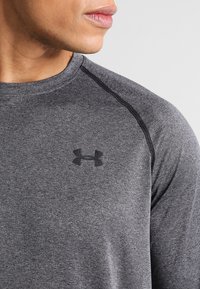 Graues Sportshirt mit rundem Halsausschnitt, das schwarze Nähte und ein dezentes schwarzes Logo auf der linken Brust zeigt. Weicher, dehnbarer Stoff.