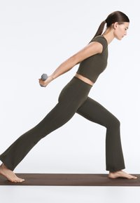 Žena v zelenom športovom oblečení vykonáva jogový výpad, pričom za chrbtom drží sivú činku na hnedom cvičebnom podložke.