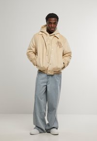 Bež suknja jakna sa kopčama na mehanizam, s logotipom na prsima, složena preko bež dukserice. Uparena sa svijetloplavim širokim trapericama i bijelim tenisicama.