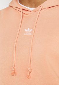 Meruňková mikina s klokaní kapsou, nastavitelným stahovacím šňůrkem a bílým logem Adidas. Měkký bavlněný materiál s hladkou texturou.