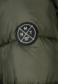 Zeleno puhvasta jakna iz tkanine z gladko teksturo; na levem ramenu se nahaja okrogla črna oznaka z belim napisom "MT 97".