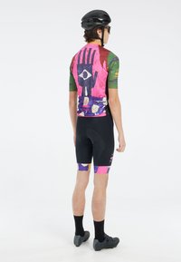 Korte mouw fietsjersey met abstracte multikleurige kunst, voornamelijk in roze en groen, gecombineerd met zwarte gepadde shorts en fietsschoenen.