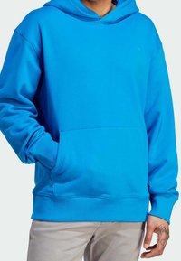 Man med ljusblå huvtröja med framficka och beige byxor, hand med flammtatuering synlig nära fickan.