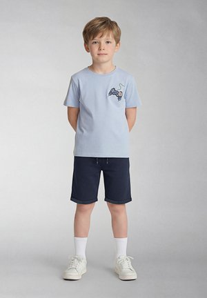 Garçon debout, mains derrière le dos, portant un t-shirt bleu clair avec un motif de manette de jeu vidéo, un short bleu marine, des chaussettes blanches et des baskets.