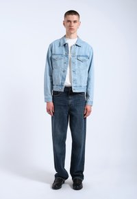 Chaqueta de denim azul claro con dos bolsillos en el pecho, llevada sobre una camisa blanca, combinada con unos jeans de pierna ancha azul oscuro y zapatos negros.