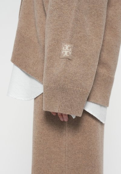 Beige pletený svetr s rukávy a sukní, na rameni s bílým logem, přes bílou žebrovanou manžetu trička; ruka částečně viditelná pod rukávem.