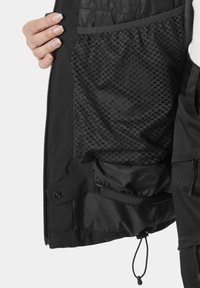 Wnętrze czarnej kurtki zawiera siatkową kieszeń z okrągłymi wzorami, gładką podszewkę, elastyczne detale oraz sznurek ściągający na dole.