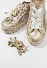 Zapatillas plataforma doradas con parte superior metálica texturizada, cierre de cordones en la parte delantera y decoración floral. Incluye un accesorio floral a juego.