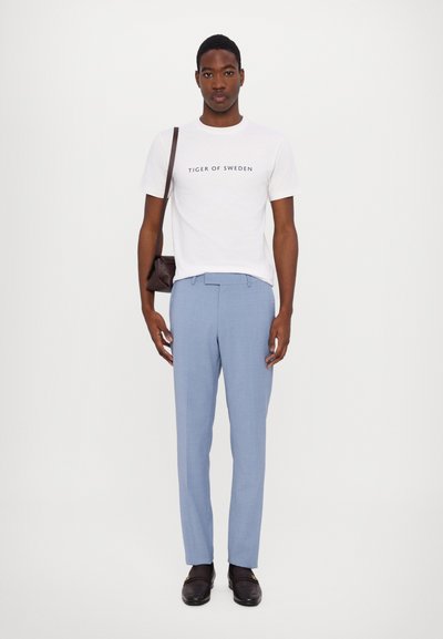 Weißes T-Shirt mit dem Text "TIGER OF SWEDEN", hellblaue maßgeschneiderte Hose, braune Lederloafers und eine braune Umhängetasche.