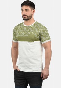 Grøn og hvid T-shirt med korte ærmer, præget af et palmebladsmønster på den øverste del, med en brystlomme og en ensfarvet hvid nederste del.